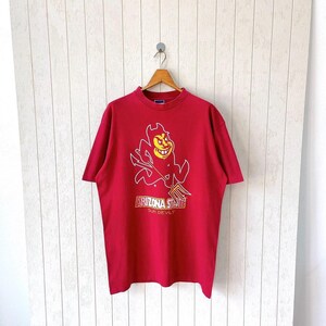 May include: A red t-shirt featuring a cartoon devil with a pitchfork, outlined in white and yellow. The text "ARIZONA STATE SUN DEVILS" is printed below the graphic in gold lettering. The shirt is hanging on a wooden hanger.