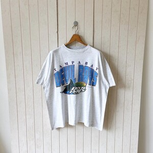 Könnte beinhalten: Ein hellgraues kurzärmeliges T-Shirt mit dem Schriftzug TAMPA BAY DEVIL RAYS in blauen Buchstaben. Unter dem Text befindet sich eine Grafik eines schwarzen Stachelrochens, der über einem regenbogenfarbenen Bogen schwimmt, mit dem Schriftzug DEVIL RAYS in Weiß und Schwarz.