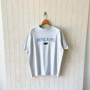 Peut inclure: Un t-shirt gris chiné avec l'inscription "DEVIL RAYS" en bleu. Le t-shirt présente un petit graphisme d'une raie stylisée en vert et noir. Le t-shirt est suspendu à un cintre en bois contre un mur de couleur claire.
