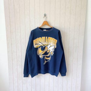 Könnte beinhalten: Marineblaues Sweatshirt mit dem Schriftzug "GEORGIA TECH" in Gold über einer Cartoon-Biene in Gelb und Schwarz mit weißen Flügeln. Das Sweatshirt hängt an einem Holzbügel.