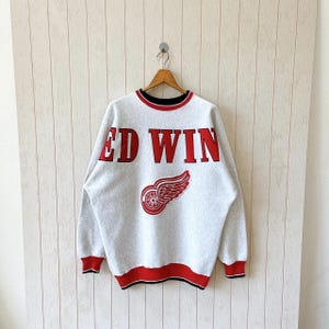 Könnte beinhalten: Hellgraues Sweatshirt mit dem Schriftzug "RED WINGS" in roten Buchstaben auf der Brust. Darunter befindet sich ein rot-weißes Logo eines geflügelten Rades. Kragen und Bündchen sind rot mit schwarzem Besatz.