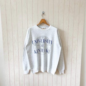 May include: A light gray crewneck sweatshirt with the words "UNIVERSITY OF KENTUCKY" in blue lettering. The sweatshirt is hanging on a wooden hanger against a light-colored wall.