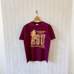 Op de afbeelding: Bordeauxrood T-shirt met korte mouwen met een afbeelding van het Arizona State University-mascotte, een duivel, en de tekst "ARIZONA STATE ASU SUN DEVILS" in goud en wit. Het shirt hangt aan een houten hanger.