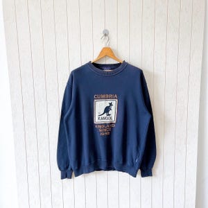 May include: Navy blue sweatshirt featuring the text "CUMBRIA" above a kangaroo logo and the text "KANGOL ENGLAND SINCE 1938". The sweatshirt is suspended on a wooden hanger against a white backdrop.