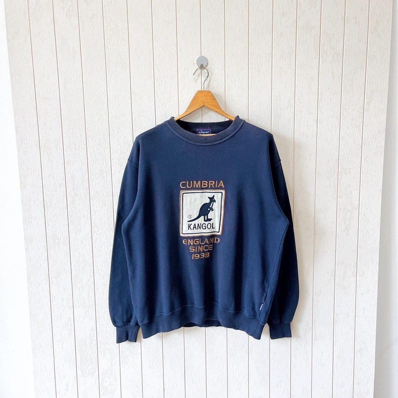 May include: Navy blue sweatshirt featuring the text "CUMBRIA" above a kangaroo logo and the text "KANGOL ENGLAND SINCE 1938". The sweatshirt is suspended on a wooden hanger against a white backdrop.