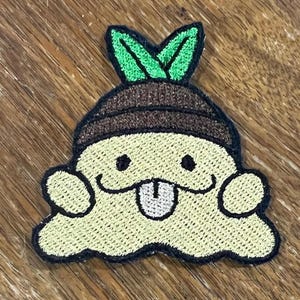 May include: An embroidered patch featuring a pale yellow, blob-shaped character with two small black eyes and a white tongue sticking out. The character wears a brown cap with two bright green leaves on top, all outlined in thick black thread.