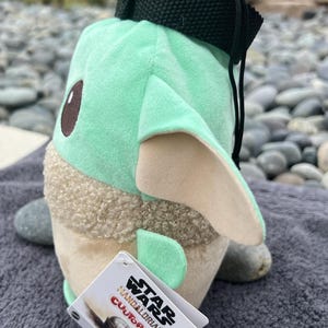 May include: A green and brown plush toy shaped like a character from the Star Wars franchise. The toy has a black strap and a tag that reads "Star Wars Mandalorian,  Cutopa, 5+".
