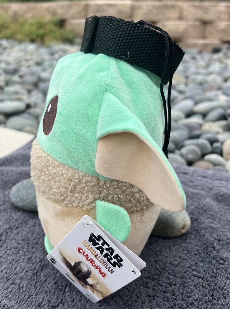 May include: A green and brown plush toy shaped like a character from the Star Wars franchise. The toy has a black strap and a tag that reads "Star Wars Mandalorian,  Cutopa, 5+".