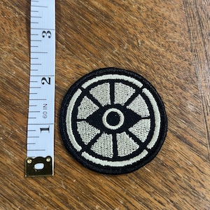 May include: A circular embroidered patch featuring a black background with a white geometric wheel-like design consisting of a central circle and six radiating segments. A white measuring tape is placed alongside, showing the patch is 2 inches wide.