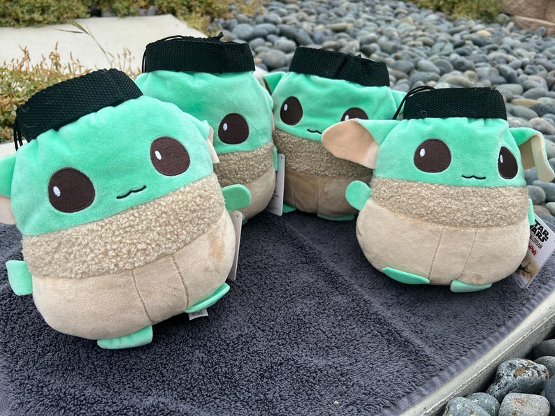 May include: Four plush green and brown Baby Yoda-themed drawstring bags. The bags have a fuzzy texture and are shaped like the character from the Star Wars series.