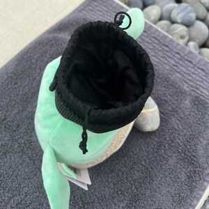 May include: A light green plush toy with a black drawstring pouch inside. The toy has a small, white, fluffy tail and a small, white, fluffy ear.