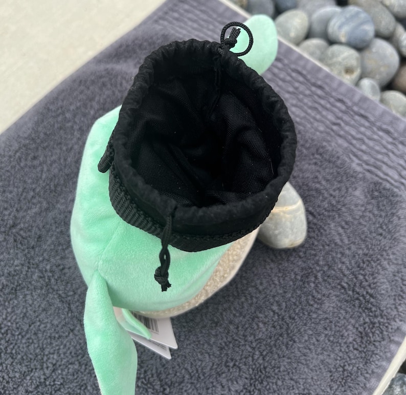 May include: A light green plush toy with a black drawstring pouch inside. The toy has a small, white, fluffy tail and a small, white, fluffy ear.