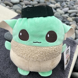 May include: A green and brown plush toy shaped like a character from the Star Wars franchise. The toy has a black strap on its head and the text "Star Wars" and "The Child" on the tag.