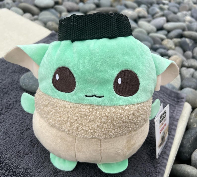 May include: A green and brown plush toy shaped like a character from the Star Wars franchise. The toy has a black strap on its head and the text "Star Wars" and "The Child" on the tag.