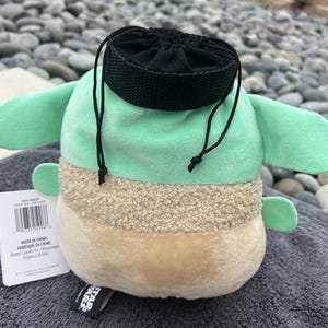 May include: A green and brown plush toy shaped like a character from Star Wars. The toy has a black drawstring bag on its head. The toy has a tag that says "Made in China. Fabriqu&eacute; en Chine. Mattel Canada Inc., Mississauga, Ontario, Canada L5N 3M2."