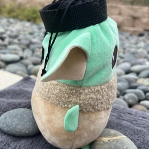 May include: A green and brown plush toy shaped like a character from the Star Wars franchise. The toy has a black drawstring bag on top.