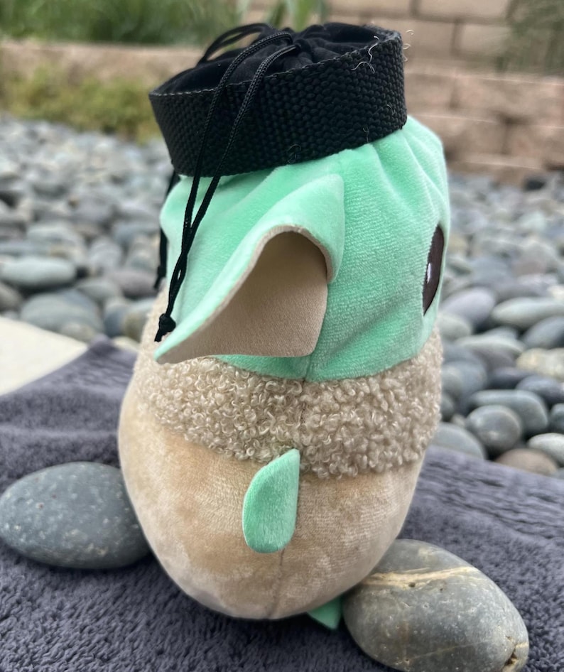 May include: A green and brown plush toy shaped like a character from the Star Wars franchise. The toy has a black drawstring bag on top.