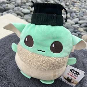 May include: Green and brown plush toy of a character from the Star Wars franchise, The Mandalorian. The toy is wearing a black fabric pouch with a drawstring. The toy is labeled "Star Wars The Mandalorian  Cufopia".