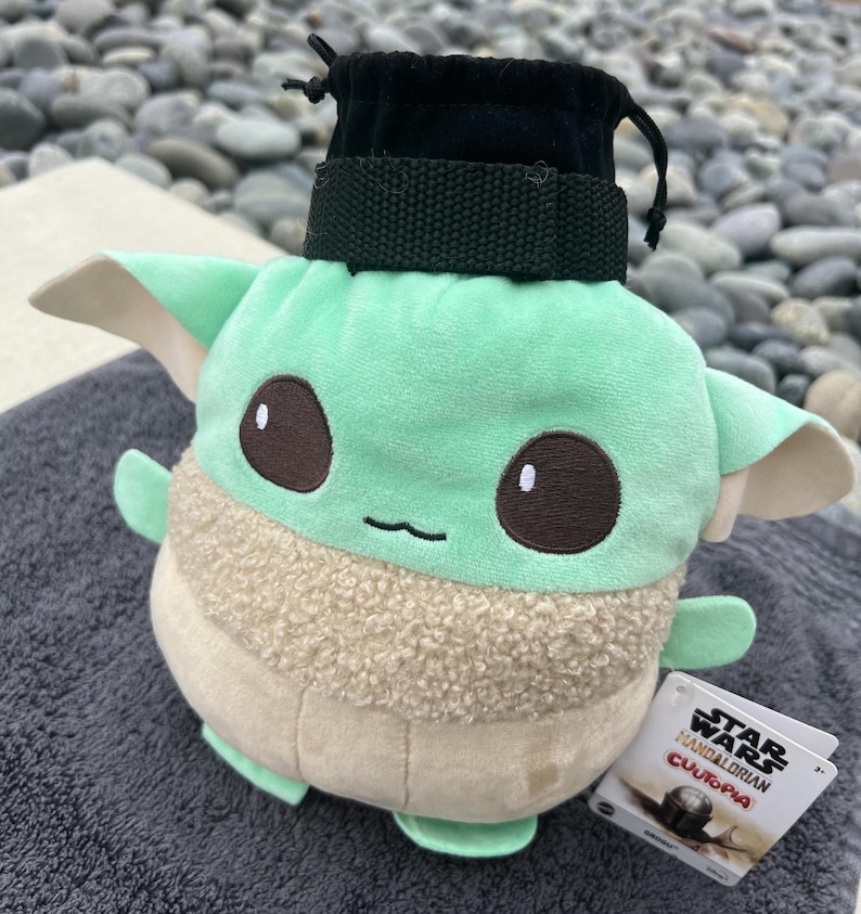 May include: Green and brown plush toy of a character from the Star Wars franchise, The Mandalorian. The toy is wearing a black fabric pouch with a drawstring. The toy is labeled "Star Wars The Mandalorian  Cufopia".