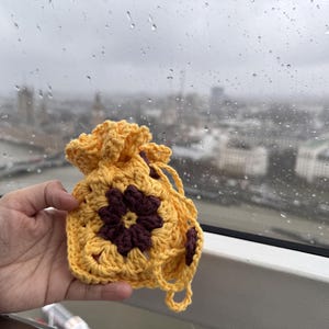 May include: A yellow crocheted drawstring bag with a brown flower design. The bag is held in a hand against a blurry background of a city.