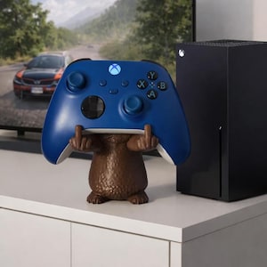 Könnte beinhalten: Ein blauer Xbox-Controller liegt auf einer braunen Figur mit erhobenen Mittelfingern. Der Controller hat schwarze Tasten und ein leuchtendes Xbox-Logo. Rechts befindet sich eine schwarze Xbox Series X-Konsole.