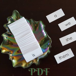 Peut inclure: Une pile de fiches blanches dans un plat vert iridescent avec les mots "is", "a", "we", "she", et "them" écrits sur des fiches blanches séparées.