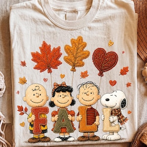 Pode incluir: Uma t-shirt cor de creme com um gráfico de Charlie Brown, Lucy, Linus e Snoopy segurando letras que formam a palavra FALL. Acima, três grandes balões em forma de folha em laranja, amarelo e castanho, rodeados por pequenas folhas de outono.