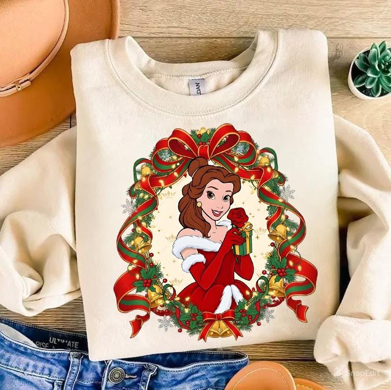May include: Cream-colored sweatshirt featuring a festive illustration of a woman in a red dress holding a rose and a gift. The design is framed by a Christmas wreath with red ribbons, bells, and greenery. The sweatshirt has a crew neck.