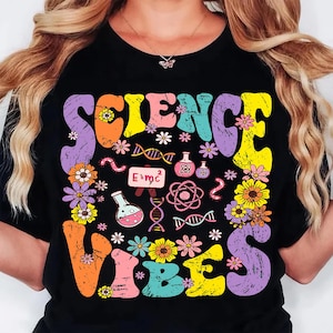 Puede incluir: Camiseta negra con las palabras "SCIENCE VIBES" en letras coloridas de estilo retro. El dise&ntilde;o incluye flores, gr&aacute;ficos tem&aacute;ticos de ciencia y la ecuaci&oacute;n E=mc&sup2;.