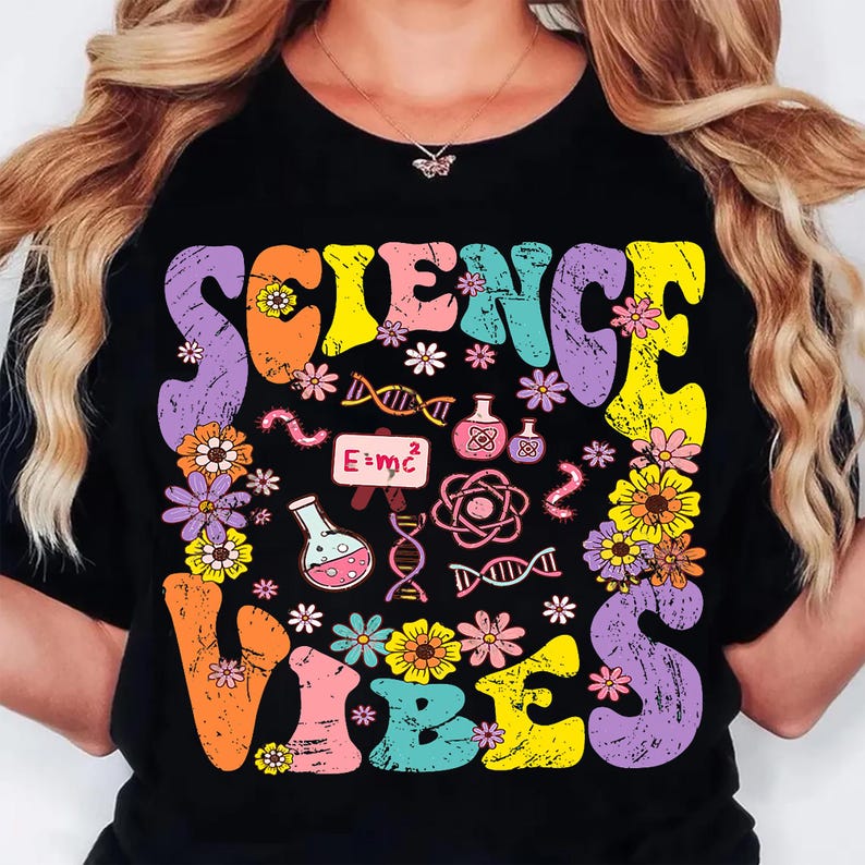 Puede incluir: Camiseta negra con las palabras "SCIENCE VIBES" en letras coloridas de estilo retro. El dise&ntilde;o incluye flores, gr&aacute;ficos tem&aacute;ticos de ciencia y la ecuaci&oacute;n E=mc&sup2;.