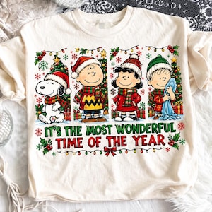 Pode incluir: Uma t-shirt de cor creme com um gráfico do Snoopy, Charlie Brown, Lucy e Linus a usar gorros e cachecóis de inverno. As personagens estão em frente a árvores de Natal com o texto, "IT'S THE MOST WONDERFUL TIME OF THE YEAR."
