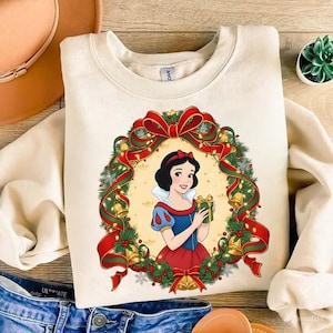 May include: Cream-colored sweatshirt featuring a graphic of Snow White holding a gift, framed by a Christmas wreath with red bows, green garland, and golden bells. A festive holiday-themed design.