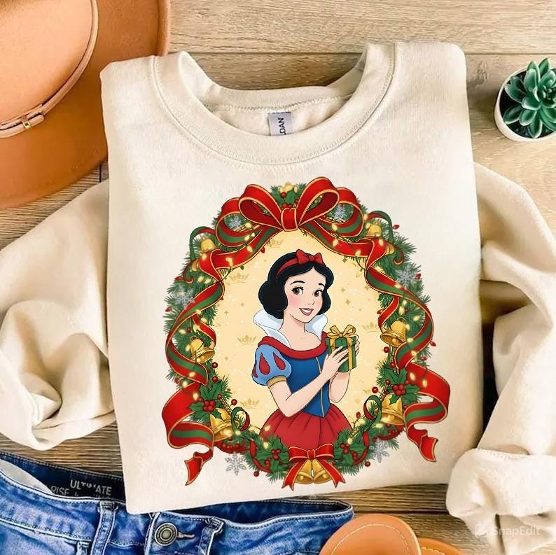 May include: Cream-colored sweatshirt featuring a graphic of Snow White holding a gift, framed by a Christmas wreath with red bows, green garland, and golden bells. A festive holiday-themed design.