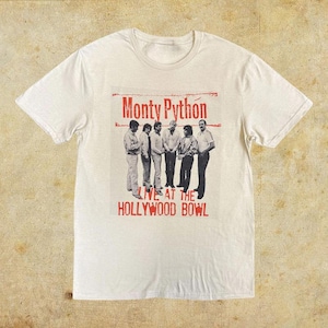 May include: Off-white t-shirt featuring a graphic of the comedy group Monty Python. The graphic includes the text "Monty Python" above a black and white image of the group, with "Live at the Hollywood Bowl" below.