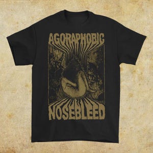 May include: Black t-shirt with the words "AGORAPHOBIC NOSEBLEED" in a distressed, gold font. The shirt features a gold illustration of a figure in a fetal position. The design has a vintage, grunge aesthetic.