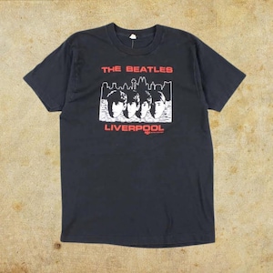 May include: Black t-shirt featuring a graphic of The Beatles with the text "THE BEATLES" above a skyline and "LIVERPOOL" below. The graphic is in white and red. The shirt is a classic crew neck style.