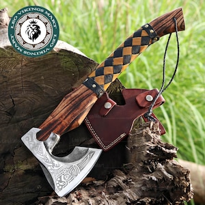 Puede incluir: Un hacha vikinga con mango de madera y empuñadura envuelta en cuero con un patrón de diamantes negros y beige. La cabeza del hacha es plateada con diseños grabados. Una funda de cuero marrón protege la hoja. El hacha se exhibe sobre una superficie de madera.