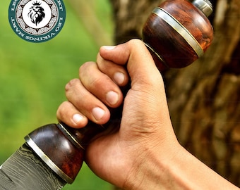 Könnte beinhalten: Nahaufnahme eines Schwertes mit Holzgriff und gemusterter Klinge. Der Griff ist dunkelbraun mit silbernen Akzenten. Die Klinge hat ein detailliertes, strukturiertes Muster. Das Schwert wird in einer Hand gehalten.