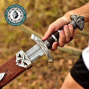 Puede incluir: Imagen detallada de una espada vikinga con una hoja estampada y una guarda y empuñadura ornamentadas de color plateado. El mango está envuelto en cuero marrón oscuro. La espada está parcialmente enfundada en una vaina de madera con detalles metálicos plateados.