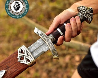 May include: A detailed image of a Viking sword with a patterned blade and ornate silver-colored crossguard and hilt. The handle is wrapped in dark brown leather. The sword is partially sheathed in a wooden scabbard with silver-colored accents.