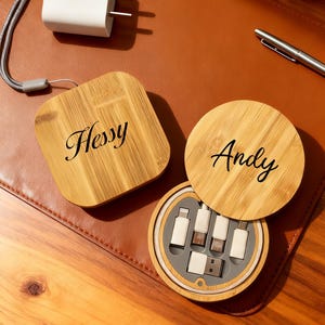 Puede incluir: Un cargador USB blanco y dos organizadores de cables de madera. Un organizador es cuadrado y dice "Hessy", el otro es redondo y dice "Andy". El organizador redondo está abierto, revelando varios cables de carga blancos.