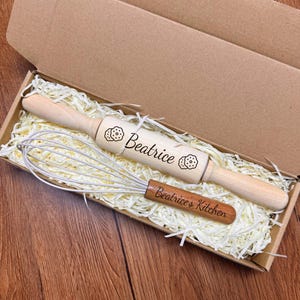 May include: A light-colored wooden rolling pin engraved with the name Beatrice and cookie icons, paired with a wire whisk featuring a wooden handle engraved with Beatrice's Kitchen, both nestled in a cardboard gift box with shredded paper filler.