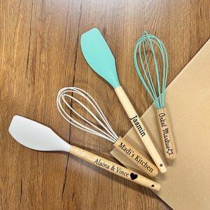 May include: Four kitchen utensils: two spatulas and two whisks. The spatulas have white and teal silicone heads with wooden handles. The whisks have white and teal wire heads with wooden handles. The handles are engraved with names and phrases.