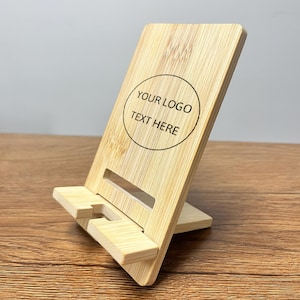 May include: A bamboo phone stand with a rectangular back and a small base. The stand has a slot to hold a phone and features the text "YOUR LOGO TEXT HERE" inside a circle.