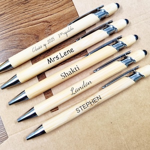 Può includere: Un gruppo di penne in bambù personalizzate con dettagli argentati. Ogni penna è incisa con un nome o un messaggio, tra cui "Class of 2025 Magalda", "Mrs. Lene", "Shakti", "Landon" e "STEPHEN". Sono disposte su una superficie marrone.