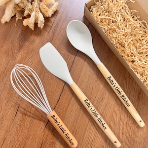 May include: A white silicone whisk, spatula, and spoon set with wooden handles. The handles are engraved with "Belta's Little Kitchen". The set is displayed on a wooden surface, with a cardboard box filled with shredded paper in the background.