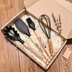 May include: A set of four black silicone kitchen utensils with light wood handles, including a pasta server, spatula, basting brush, and whisk, arranged in a cardboard gift box with shredded paper filler and a sprig of dried lavender.