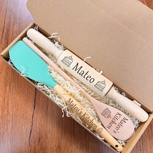 May include: A wooden rolling pin, spatula, and spoon set in a cardboard box. The rolling pin and spoon are engraved with a cake design and the name "Mateo". The spatula has a teal silicone head.