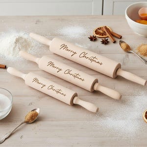 Può includere: Tre mattarelli in legno con la scritta "Merry Christmas". Sono disposti su una superficie di legno chiaro, con farina e ingredienti per la cottura sparsi intorno.