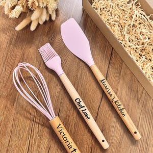 Peut inclure: Ensemble d'ustensiles de cuisine roses et en bois, comprenant un fouet, un pinceau et une spatule. Les manches en bois sont gravés des noms "Victoria", "Chef Love" et "Marissa's Kitchen". L'ensemble est présenté sur une surface en bois.
